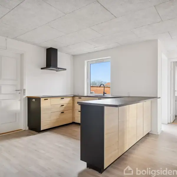 Moderne køkken med træskabe og sort bordplade, der står i et lyst rum med vindue. En dør og en gang forbinder køkkenet med andre rum.