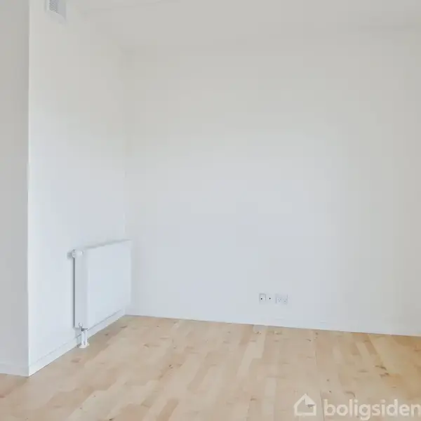 Tomt rum med hvide vægge og trægulv. En radiator er monteret på væggen til venstre. Rummet er lyst og minimalistisk, uden møbler eller dekorationer.
