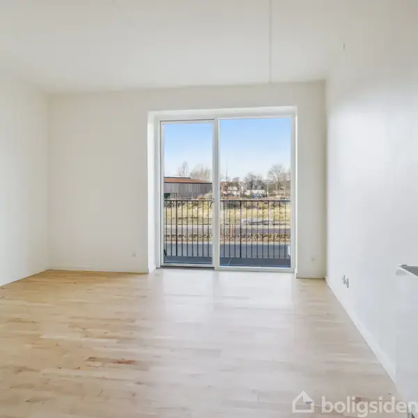Stue med lyst trægulv går ud mod en stor glasskydedør med altanudsigt. Udenfor ses et åbent område med træer og bygninger i baggrunden. Ingen tekst.