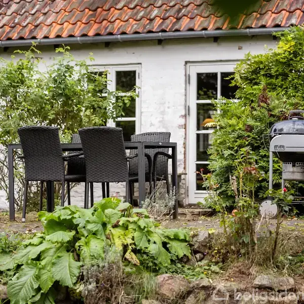 En sort havemøbelsæt står stille på en terrasse foran et hvidt murstenshus med rødt tegltag. Ved siden af er en grill, omgivet af frodig grøn beplantning.