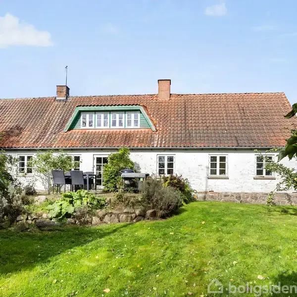 Et hvidt hus med rødt tegltag og grønne kanter har en terrasse med havemøbler foran. Det hele er omgivet af en velplejet græsplæne og bevoksning.