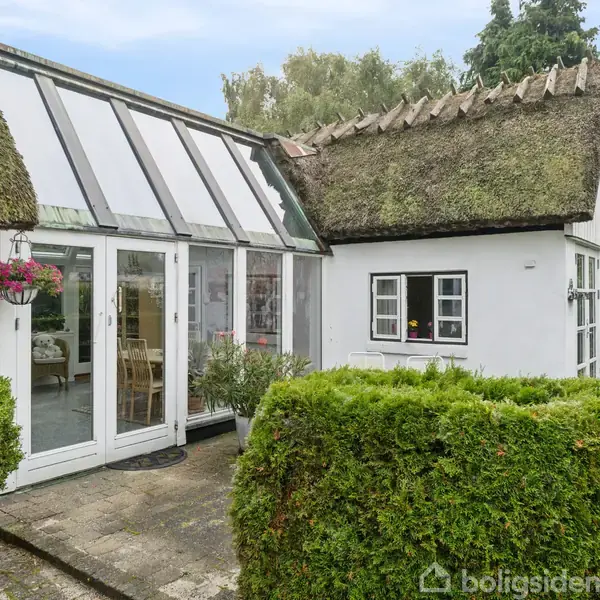 Stråtækt hus med hvide vægge har en glasudestue, som er fyldt med lys. Omkring huset er grønne buske og træer, der skaber en naturpræget ramme.