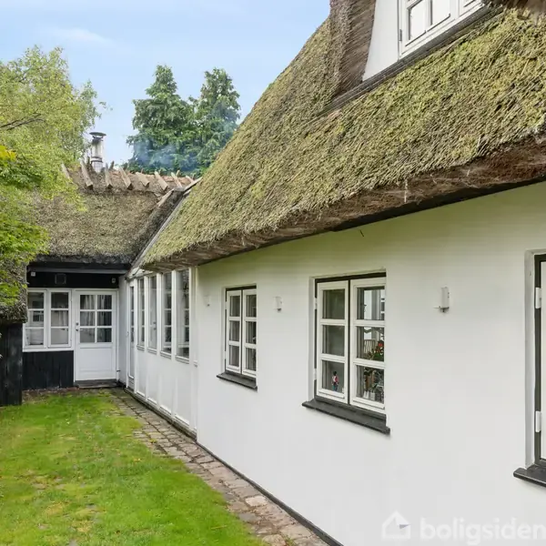 Et traditionelt hus med stråtag står stille omkranset af grønne træer og buske. Bygningen har hvide vægge og vinduer med sprosser, som skaber en idyllisk, fredfyldt stemning.