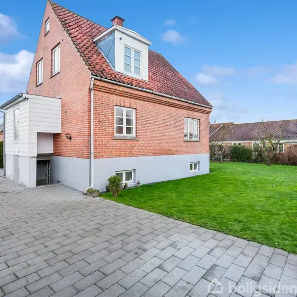 En rød murstensvilla med rødt tag står stille i en veletableret have med græsplæne og flisebelagt indkørsel, omkranset af hække og nabohuse under en blå himmel.