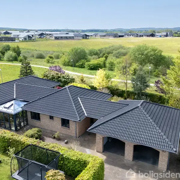 En moderne bungalow med sort tegltag og murede vægge står i et naturskønt område omgivet af grønt landskab og marker under en klar blå himmel.