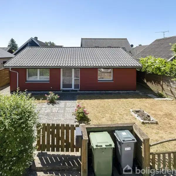 Rødt hus står stille, omgivet af en flisebelagt gårdsplads og en frodig have. Huset har et gråt tegltag og ligger i et villakvarter med flere lignende huse rundt omkring.