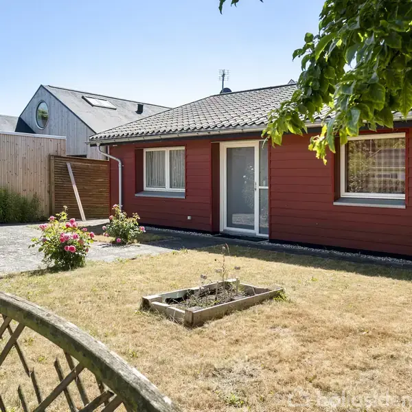 Rødt hus står på en græsplæne med blomsterbed. Foran ses en træport, og ved siden af er en grusindkørsel. Huset har et mørkegråt tag og blå hoveddør.