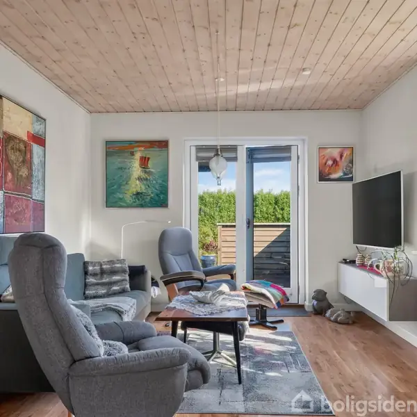 Stue med to grå lænestole og en sofa, placeret omkring et sofabord. En fladskærms-tv hænger på væggen, og rummet har træloft. Store vinduer og glasdøre åbner ud mod en terrasse. Malerier pynter væggene.