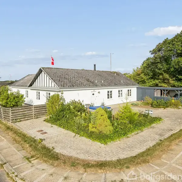Hvidt hus med sort tag og dannebrogsflag, omgivet af fliser og have med buske. Til venstre en vej og til højre træer og bilparkering.