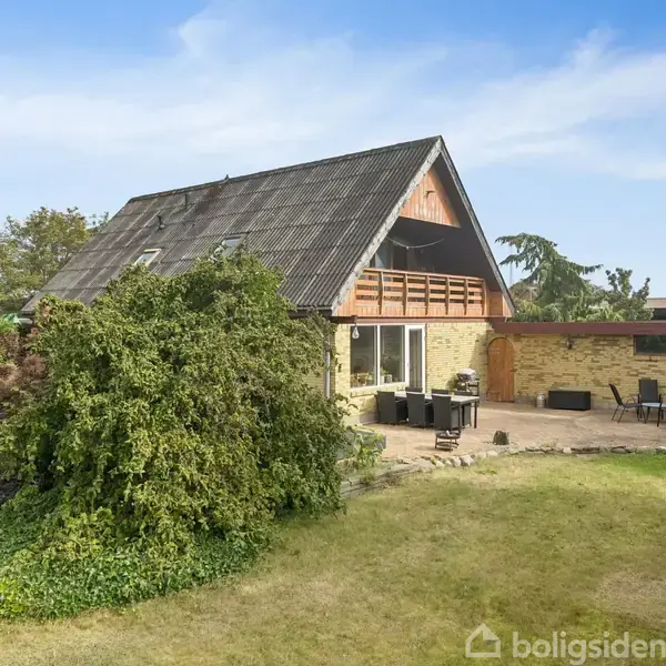 Hus med skråtag står i en have og har en stor terrasse med udendørs møbler. Omkring er der grønne buske og træer under en blå himmel.