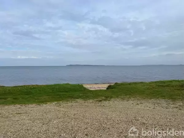 Grusvej fører til en græsplæne ved en stille kyst. I baggrunden ses en rolig havoverflade og en ø i det fjerne. Himlen er delvist skyet.