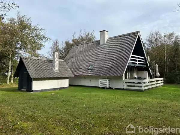 Et hvidt hus med trekantet tag står på en grøn græsplæne. Et mindre skur ses ved siden af. Træer omkranser bygningerne i et landskab.