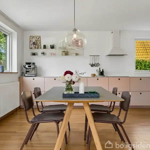 Et spisebord med blomster og lys står i et lyst køkken med hvide vægge og træpaneler. Skabe, en kaffemaskine, hylder og en emhætte er synlige i baggrunden.