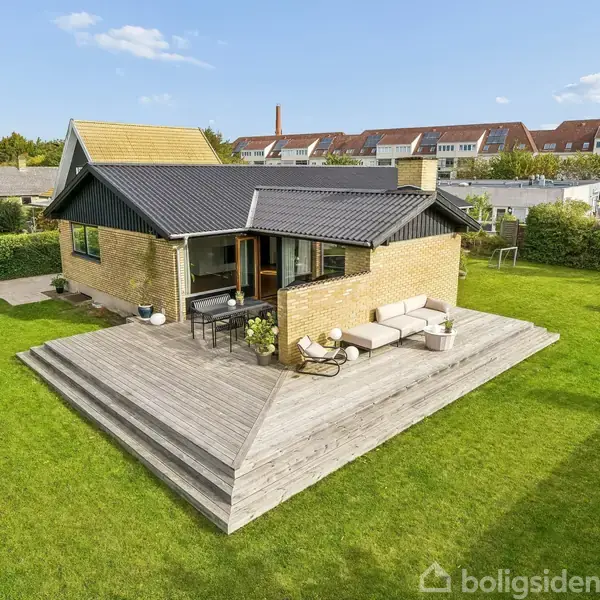 Et gulstenshus har en træterrasse med havemøbler. Huset ligger i en grøn have omgivet af hegn og nabohuse. Skyfri himmel i baggrunden skaber en solrig atmosfære.