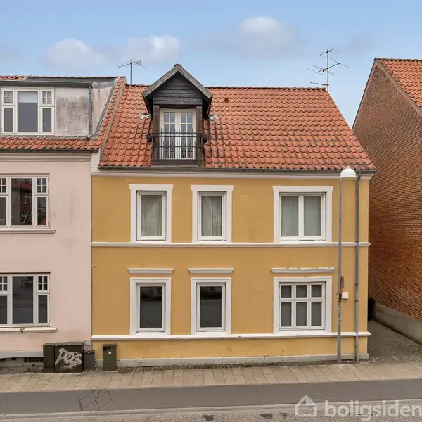 Et gult, to-etagers hus med tegltag og frontalt balkonvindue står mellem et hvidt og et rødt murstenshus. Huse ligger langs en asfalteret vej under en blå himmel.