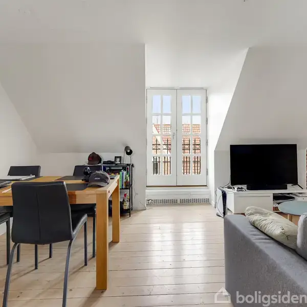 Stue med sofa og TV. Der er et spisebord til venstre med fire stole. Væggene er hvide med en eksponeret murstensvæg. Franske døre leder til en balkon.