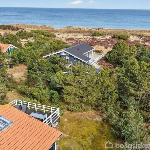 Huse omgivet af træer ligger langs en kystlinje med udsigt over havet. En bygning har en træterrasse og rødt tag, mens anden har gråt tag.