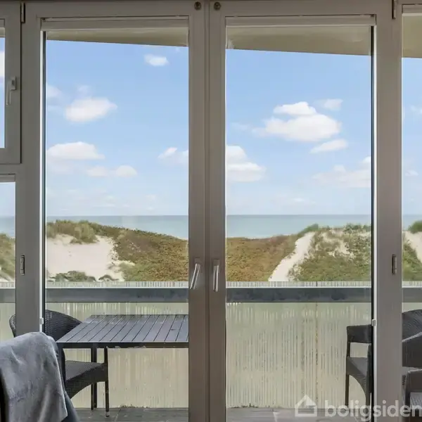 Døre fører ud til en balkon med to stole og et bord. Udsigten viser sandklitter og en blå himmel med skyer i baggrunden.