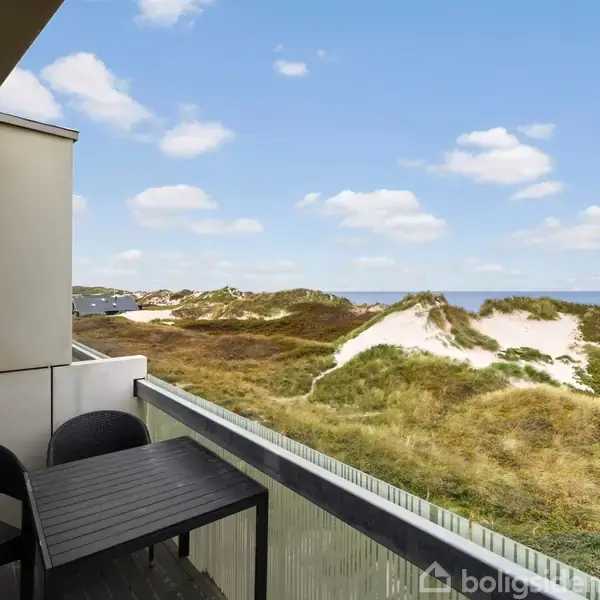 En altan med et sort bord og to stole vender mod klitter og græsarealer, med havet i baggrunden under en blå himmel med skyer.