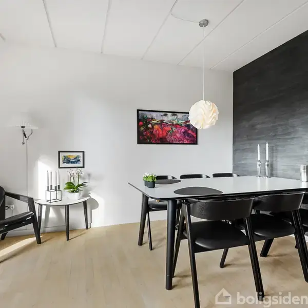 En spisestue indeholder et sort spisebord med seks stole. En pendellampe hænger over bordet, og der er kunst på væggene. Rummet har lyse vægge og trægulv.
