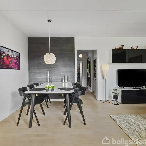 En moderne spisestue med sort bord og seks stole står i et lyst rum. På væggen hænger et maleri, og i baggrunden ses en tv-skærm og dekorative hylder.