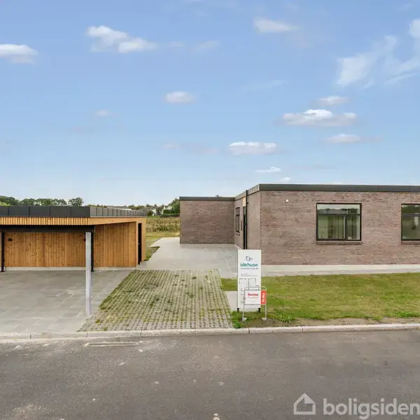 Køkken med en lang køkkenø i midten, omgivet af hvide skabe. I baggrunden ses et spisebord med røde stole nær store vinduer. Moderne, lys indretning.