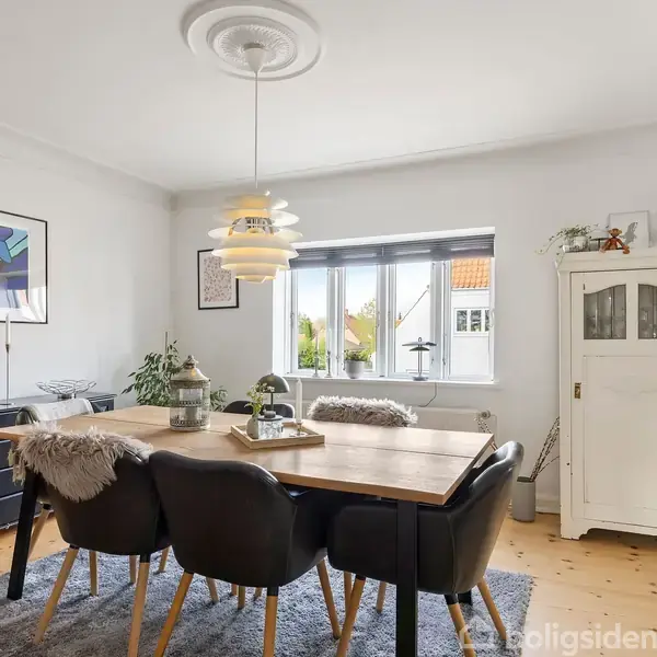 En spisebord med sorte stole står i et lyst rum med trægulv. En moderne lampe hænger over bordet, og der er kunst på væggene. Vinduer lukker lys ind.
