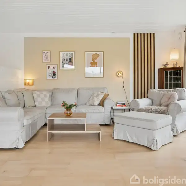 En hjørnesofa med lysegrå betræk står i en stue ved siden af en lænestol med skammel. Et lille sofabord med en plante. Væggene har dekorationer og lamper.