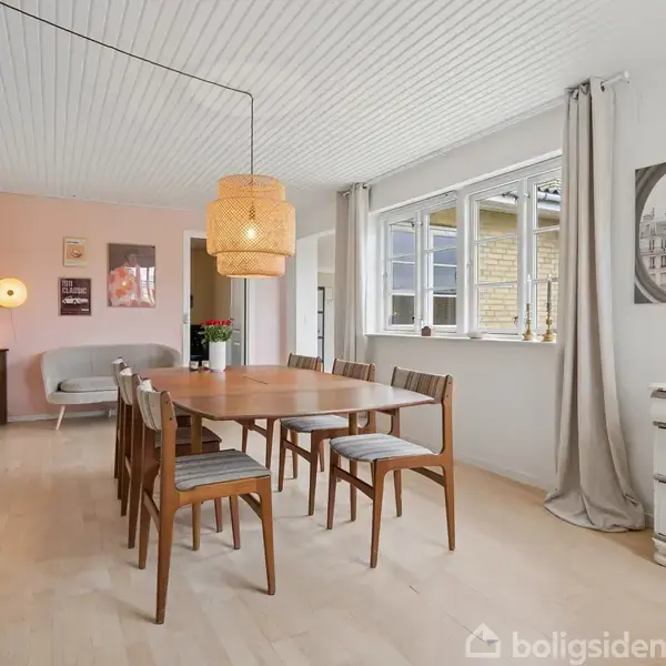 En spisestue med et træbord omgivet af otte stole. En stor lampe hænger over bordet. Væggene er lyse, rummet har et vindue, en kommode og åben dør til en stue.