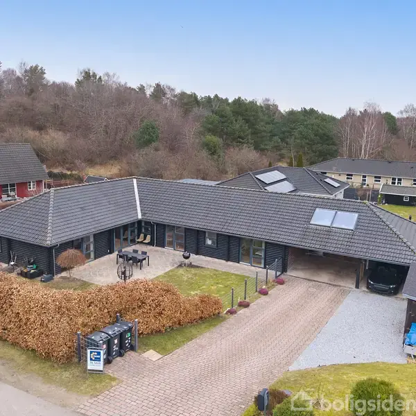 Et moderne, sort hus med grått tegltag ligger ved en sti omgivet af træer og hække. Der står cykler og havemøbler på terrassen, og en indkørsel fører til en garage.