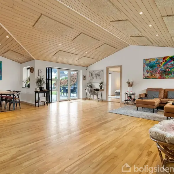 Et rummeligt stueinteriør viser et træloft og gulv. Et spisebord står til venstre, mens en sofagruppe er til højre. Store glasdøre leder til en udendørs terrasse.