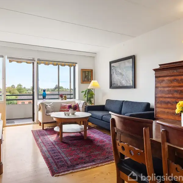 Stue med en blå sofa og træmøbler, placeret på et rødt tæppe. Et sofabord og en reol er til stede. Store vinduer giver udsigt til balkon og grøn udsigt udenfor.