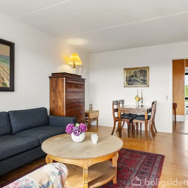 En stue med en mørk sofa, hvor der står et rundt bord med blomster foran. Der er malerier på væggene, og et spisebord med stole i baggrunden.