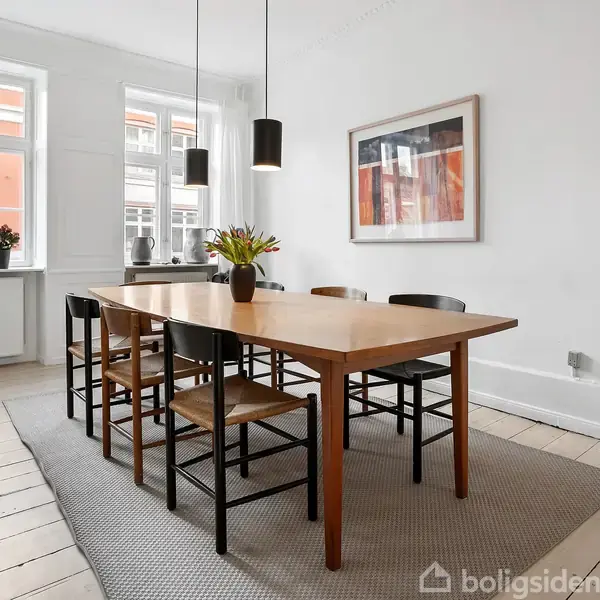 Et spisebord med seks stole står på et tæppe. Vasen med blomster er på bordet. Omgivet af hvide vægge, vinduer og vægkunst. Lamper hænger over bordet.