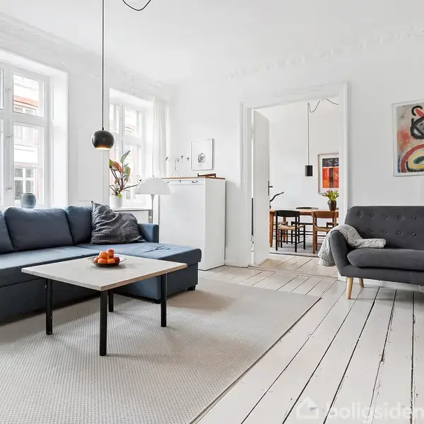 En stue med en blå sofa og sofabord står på et lyst trægulv. Et farverigt maleri hænger på væggen, og en stor reol fyldt med bøger ses til højre. I baggrunden er der adgang til et spiseområde.