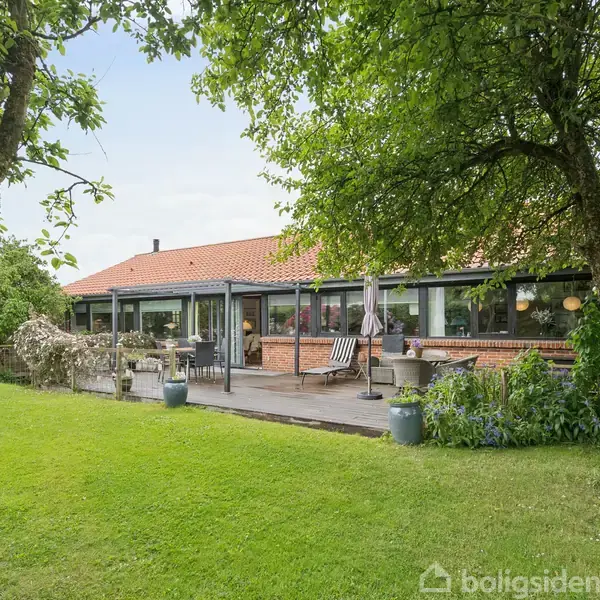 Huset er en étplans bygning med rødt tegltag, omgivet af en terrasse med havemøbler og en stor, grøn have med træer.