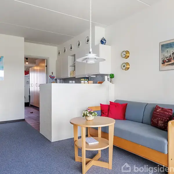Stue med en blå sofa langs højre væg, dekoreret med røde puder. Et lille rundt bord foran sofaen. Et spisebord og stole til venstre. Kunst og vægure hænger på væggene.