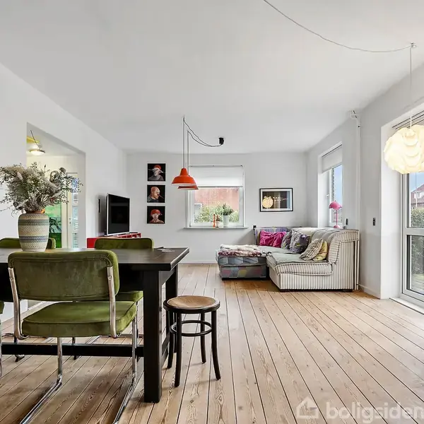 Stue med trægulv fyldt med lyse møbler; grønne stole og et stort sofabord står ved et spisebord. Vægmalerier hænger over en stor sofa ved vinduer, der fylder rummet med lys.