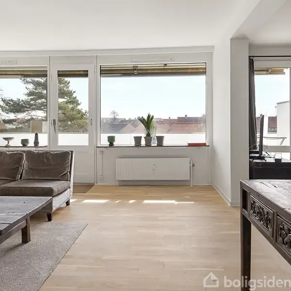 Stue med sofa, sofabord og TV; lyse trægulve og store vinduer slipper dagslys ind. Buddha-statue står på et træbord til højre. Udsigt til træer og bygninger udenfor.
