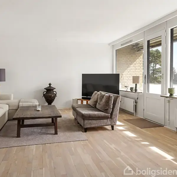 Stue med sofa og lænestol omkring et sofabord, fladskærms-TV overfor. Store vinduer og dør til altan i baggrunden. Lyse trægulve, planter og minimalistisk indretning.