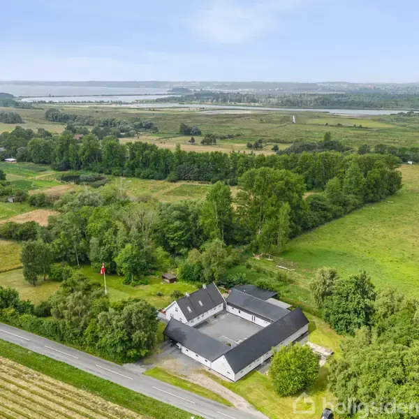 Et hvidt, vinkelformet hus ligger omgivet af træer og marker i et åbent, landligt område. I baggrunden ses en fjord. Stille atmosfære og grønne omgivelser dominerer landskabet.