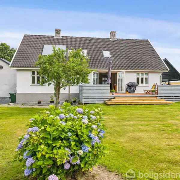 Hus med hvide vægge og skråt tag står stille. En terrasse foran huset, omgivet af frodig have med blomstrende buske og græsplæne. Træer og marker i baggrunden.