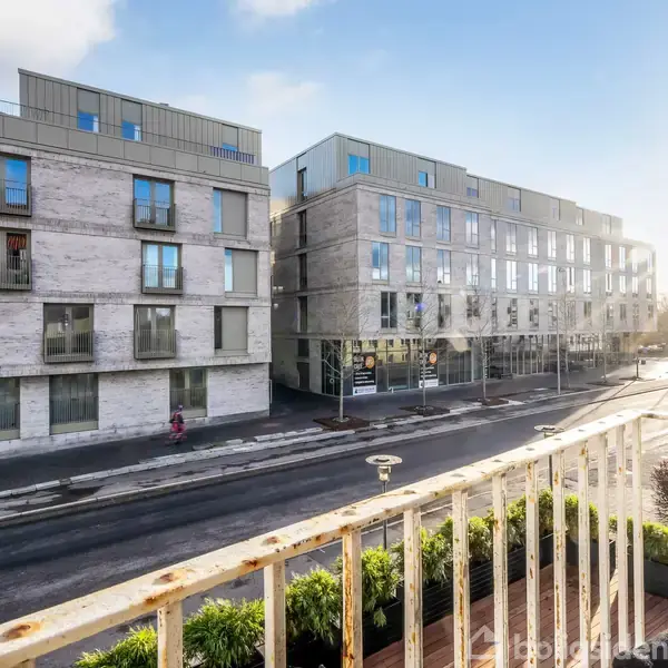 To moderne lejlighedskomplekser står parallelle på hver side af en gade. En person går på fortovet, og der er solskin. Udsigten er fra en altan med grønne planter.