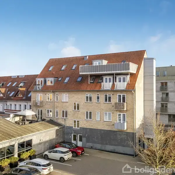 En fleretagers bygning med rødt tag står i et byområde, omgivet af andre bygninger og biler parkeret foran. En klar blå himmel ses i baggrunden.