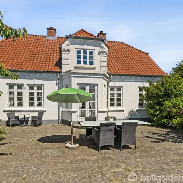 Hvid villa med rødt tegltag. Terrasse har bord, stole og grøn parasol. Omkranset af tætte buske og et stort, frodigt træ til venstre. Solrigt vejr.