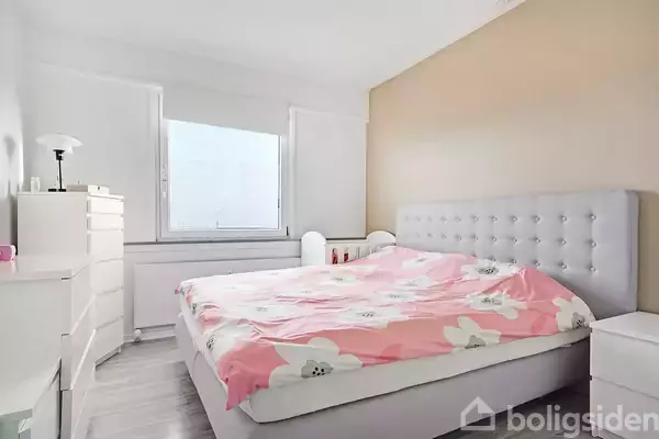 En seng med blomsterbetræk ligger i hjørnet af et lyst værelse. En kommode er placeret ved siden af vinduet, og en bordlampe står på den. Rummets vægge er delvist malet lysebrune.