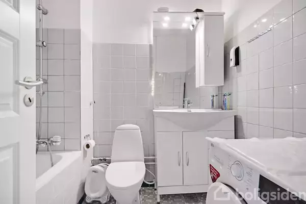 Et badeværelse med hvide fliser og moderne inventar. En toalett, håndvask med spejl og en vaskemaskine er synlige. Bruseområdet er adskilt af fliser. Rummet er lyst og enkelt.