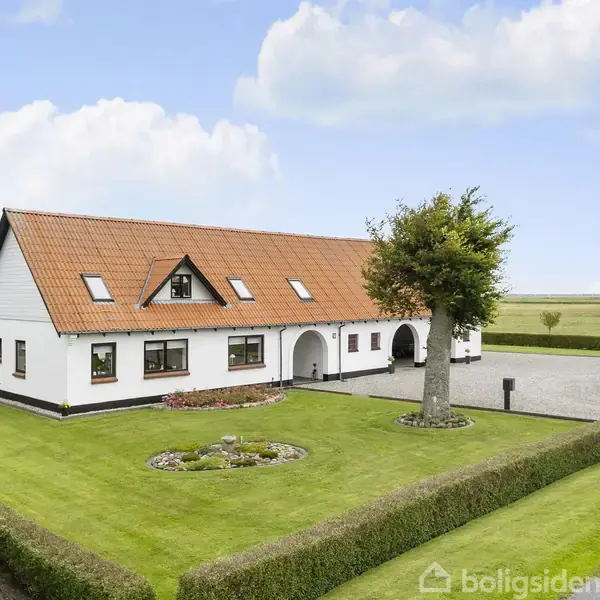 Et hvidt hus med rødt tegltag står i en veltrimmet have med grøn plæne og gamle træer, omgivet af marker og blå himmel.