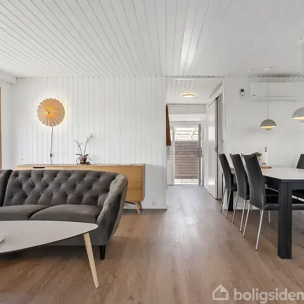 Grå sofa og sofabord står i en stue med hvide vægge og trægulv. Ved siden af er et spisebord med sorte stole. Store vinduer giver naturligt lys.