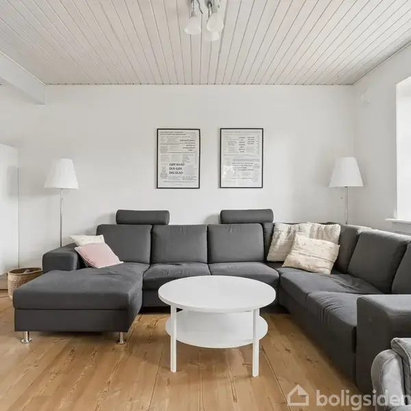 En mørkegrå hjørnesofa med puder står i en stue. Ved siden af er der en hvid rund sofabord og to gulvlamper. En hvid skænk og vindue ses i baggrunden.